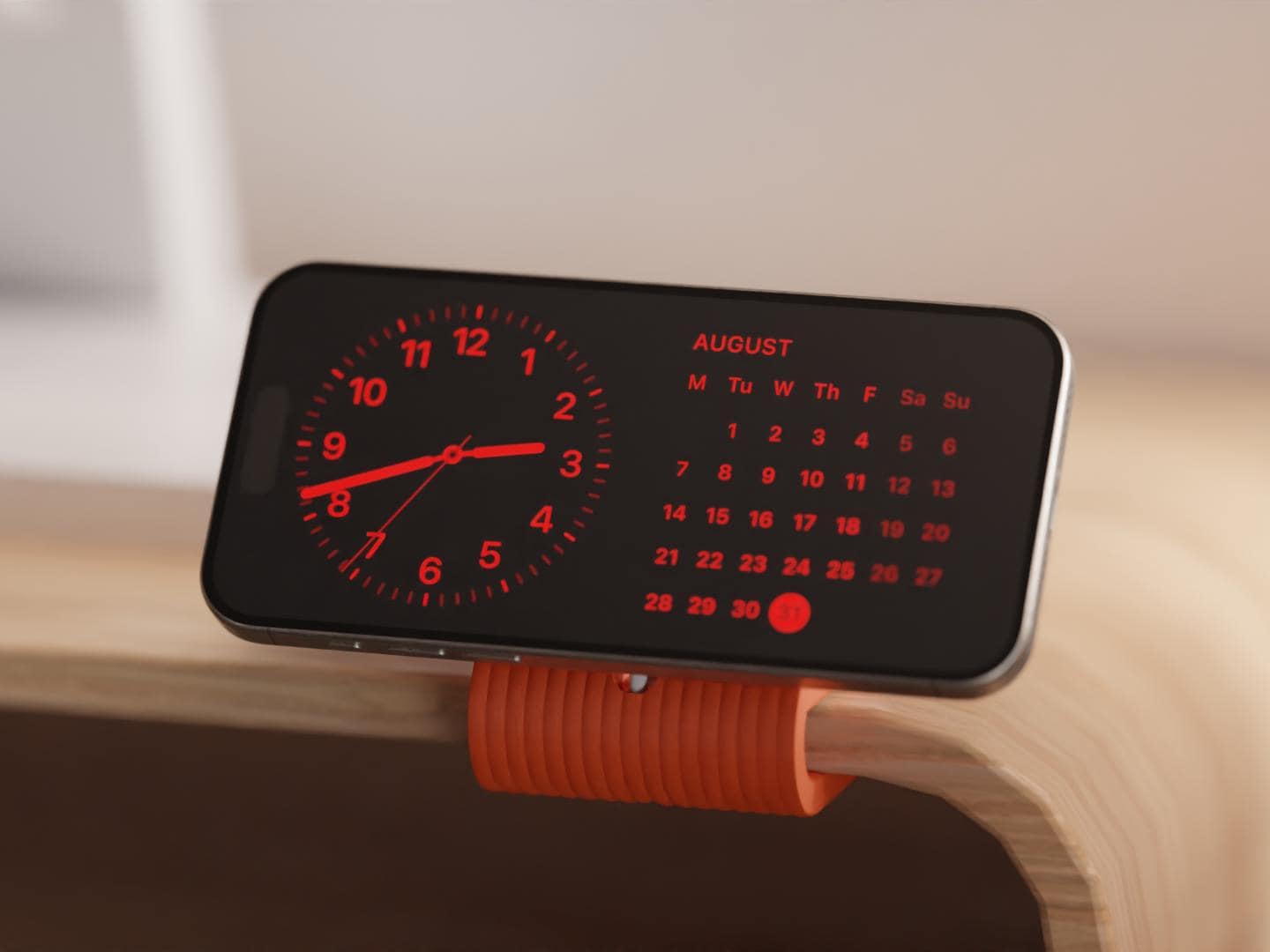 Gerippter Monitor-Clip MagSafe Halterung in leuchtendem Orange, befestigt am Rand eines Holz-Monitorständers. Das minimalistische Design hält das iPhone perfekt im StandBy Modus auf Augenhöhe.